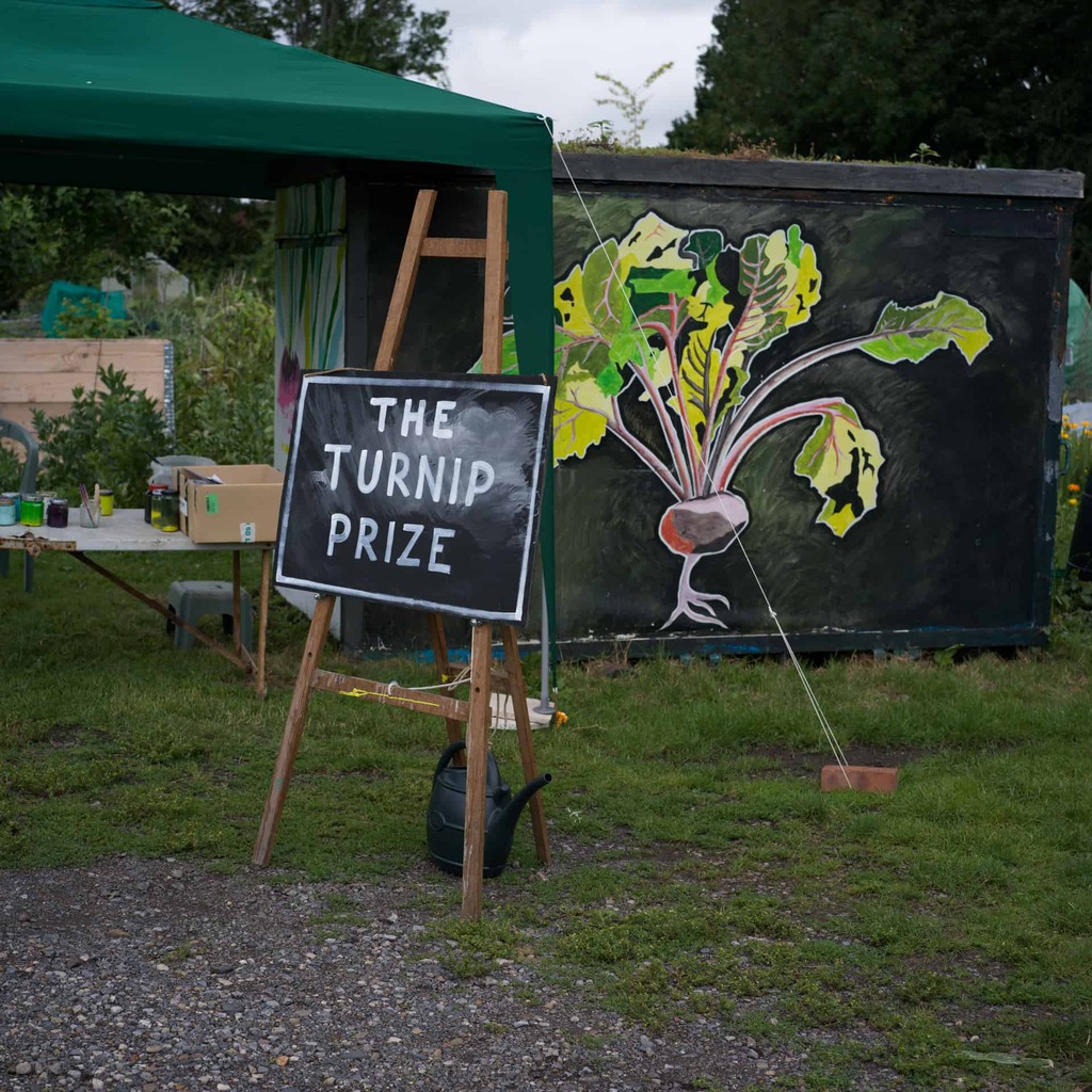 a photograph in an allotment. A sign reads, 'The Turnip Prize'. There is also a beetroot painted in the side of a shed.