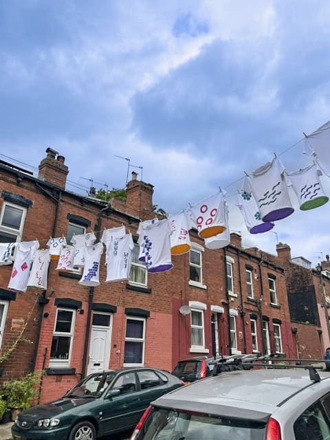 art installation. A row of t-shirts, with art work emblazoned on them, are hanging high on a washing line between two back-to-back houses.