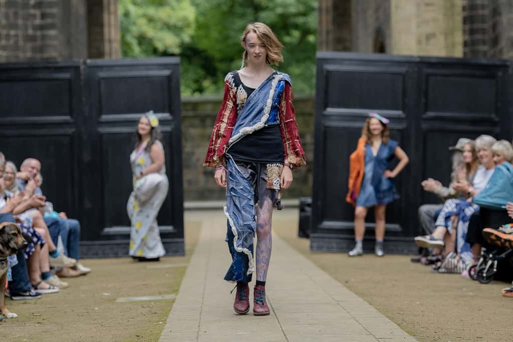 The model is wearing a top and trousers made out of many different materials and colours. One of the legs is cut short.