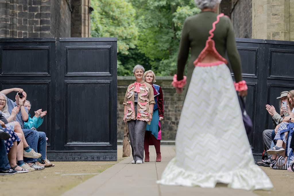 A model walks away from the camera wearing a long dress with open back. There are models waiting to walk standing in the background
