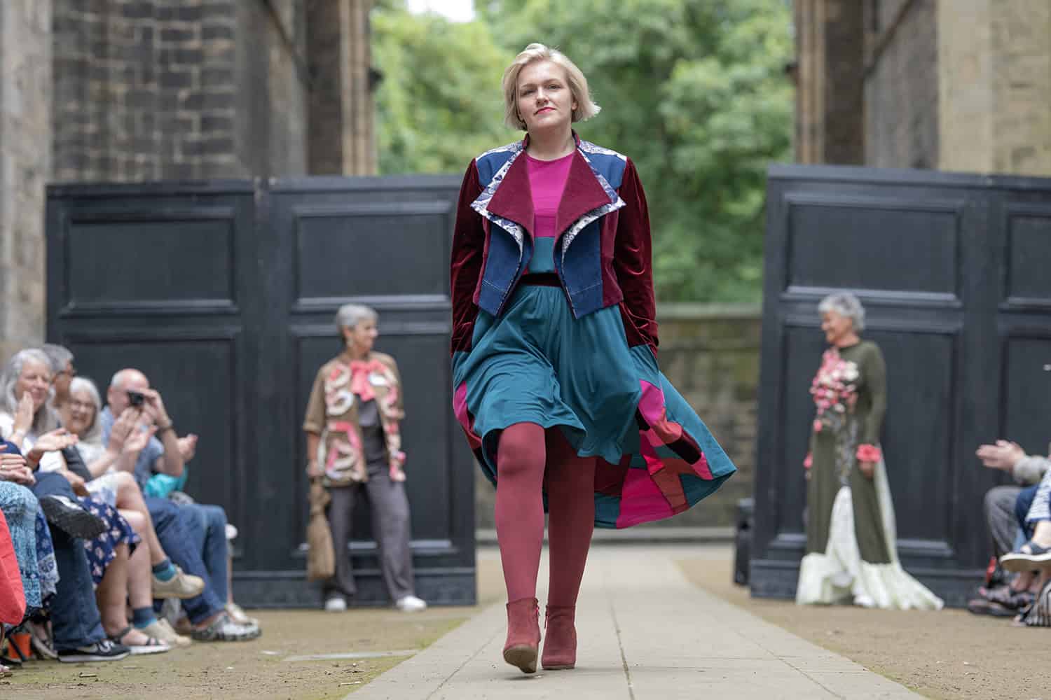 The model wears a brightly coloured top and skirt, with a velvet and denim jacket.
