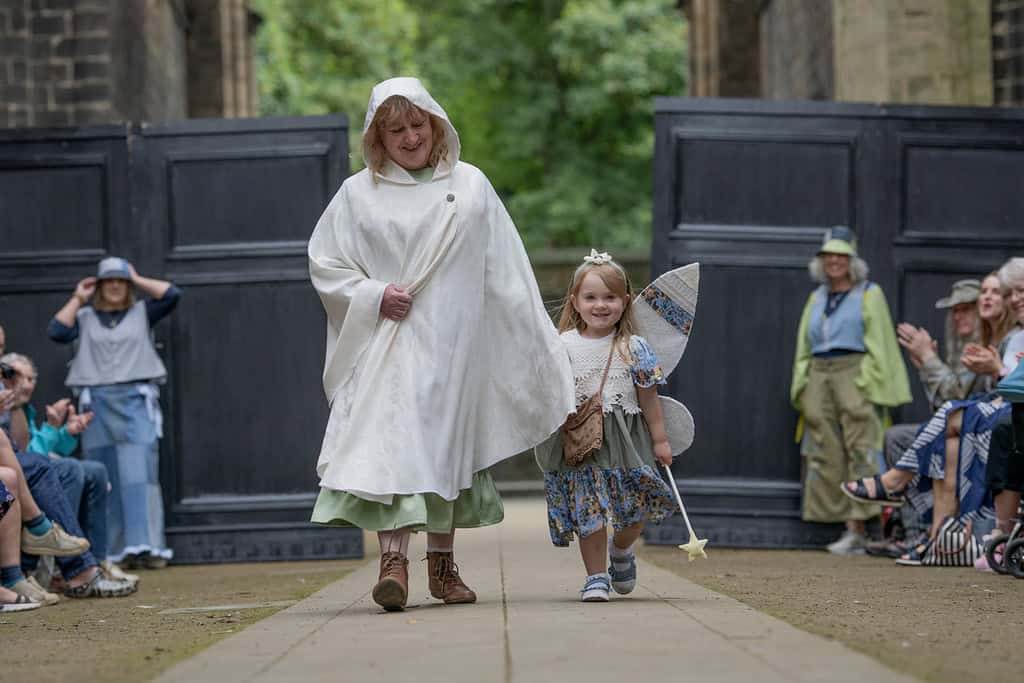 A young girl wears fairy wings and a dress. She is accompanied by an adult model who is wearing a hooded cape