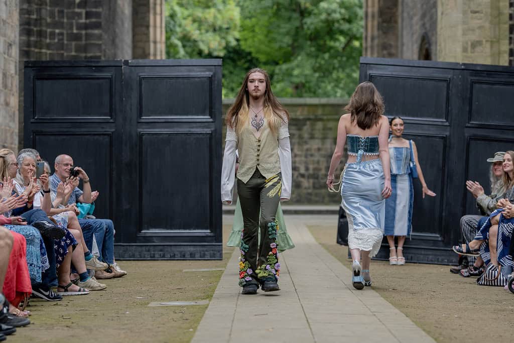 One model wears a shirt and flared trousers. A second model wears a laced up top and long skirt.