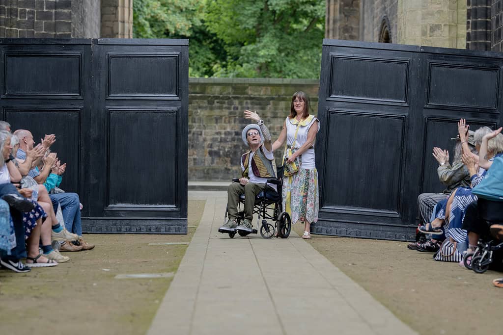 One model wears a long dress, another model and wheelchair user wears a hat, waistcoat and trousers