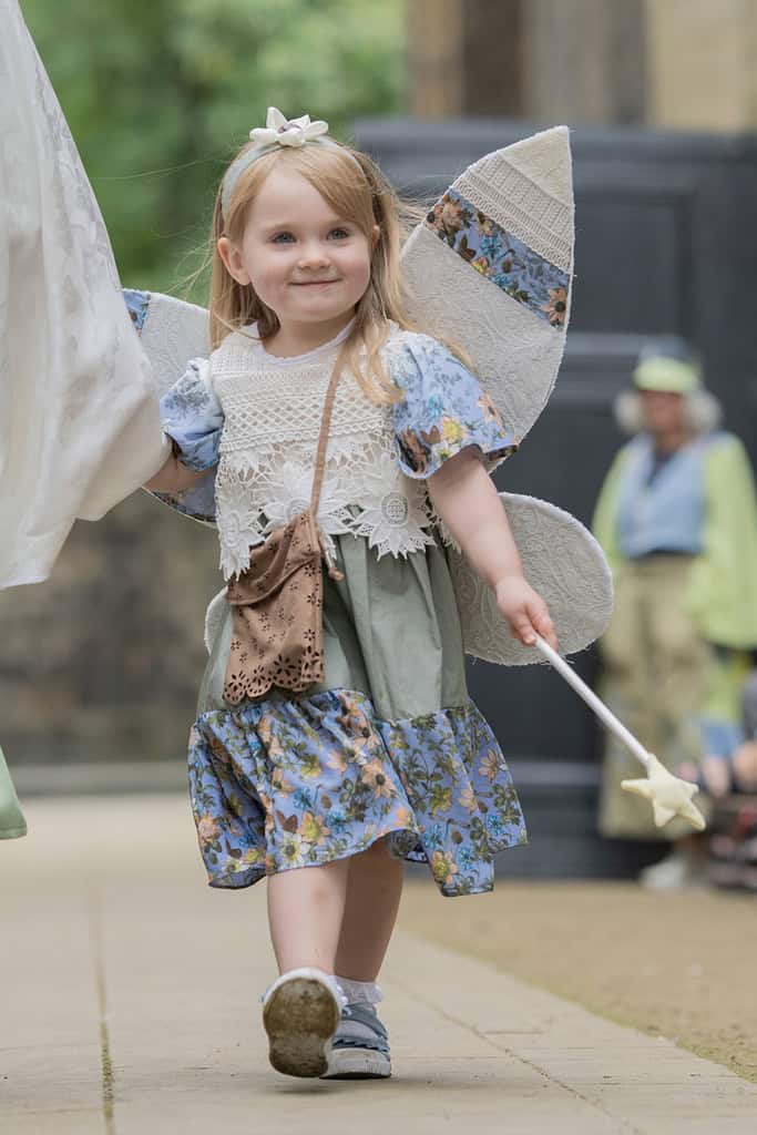 A young girl wears fairy wings and a matching dress, she carries a wand.