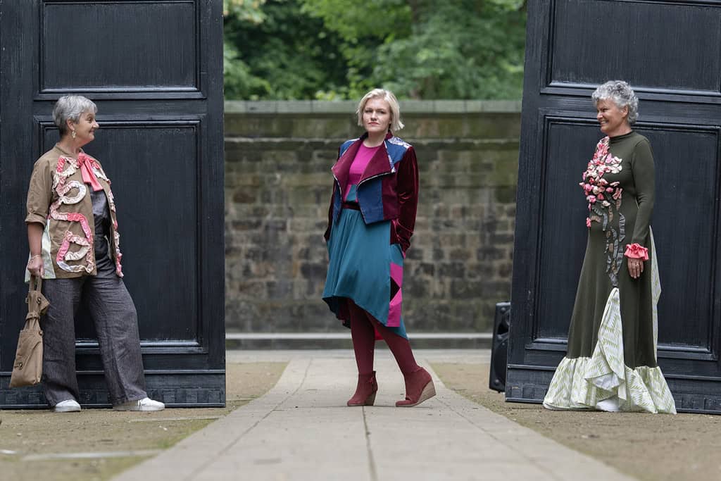 There are three models. The one on the left wears a top and trousers and a jacket decorated with appliqués. The middle one wears a colourful dress and jacket. The one on the right wears a long dress decorated with appliqués.