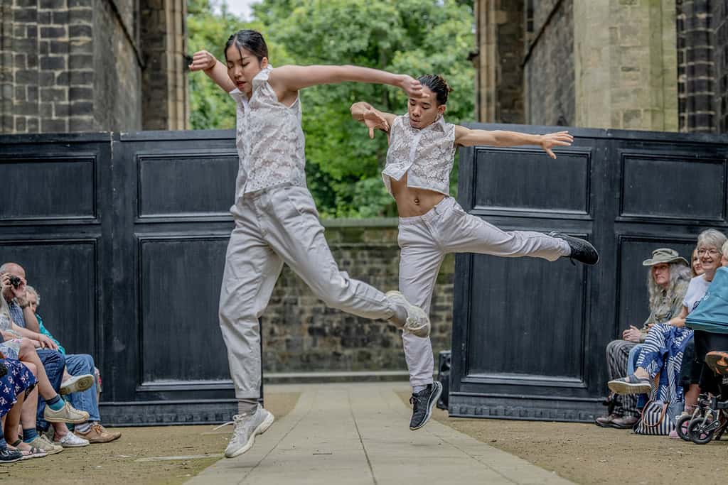 Two dancers perform, they can be seen leaping from the ground. They wear sleeveless lace shirts that were created by one of the designers.