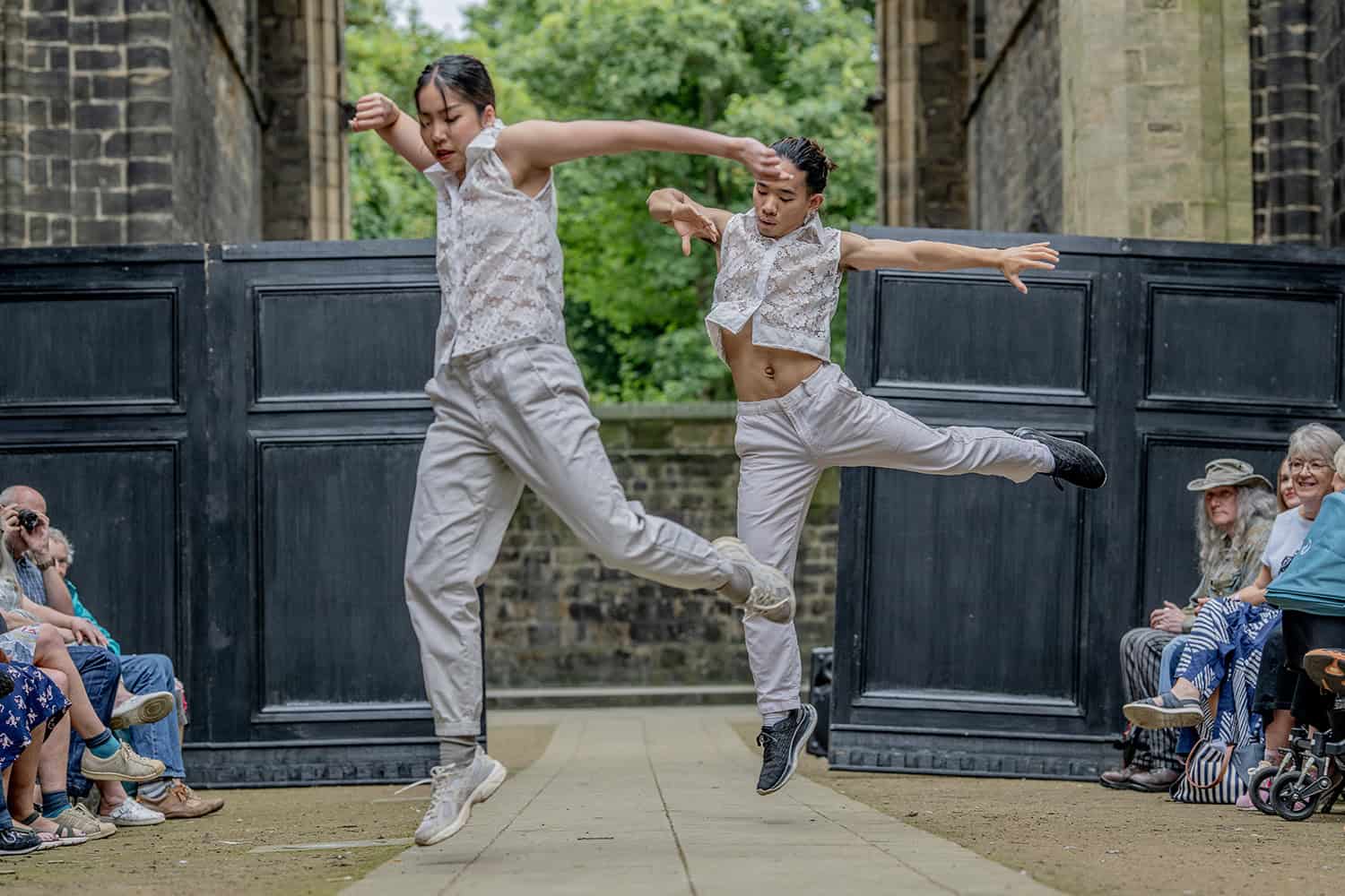 Two dancers perform, they can be seen leaping from the ground. They wear sleeveless lace shirts that were created by one of the designers.
