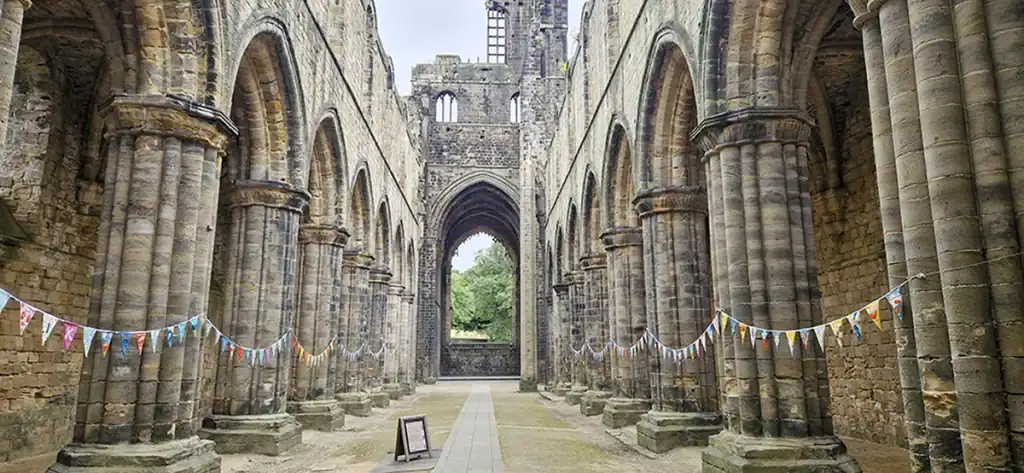 View down the ruined nave of Kirkstall Abbey Church, with rows of stone arcade columns and colourful bunting strung between them on both sides.