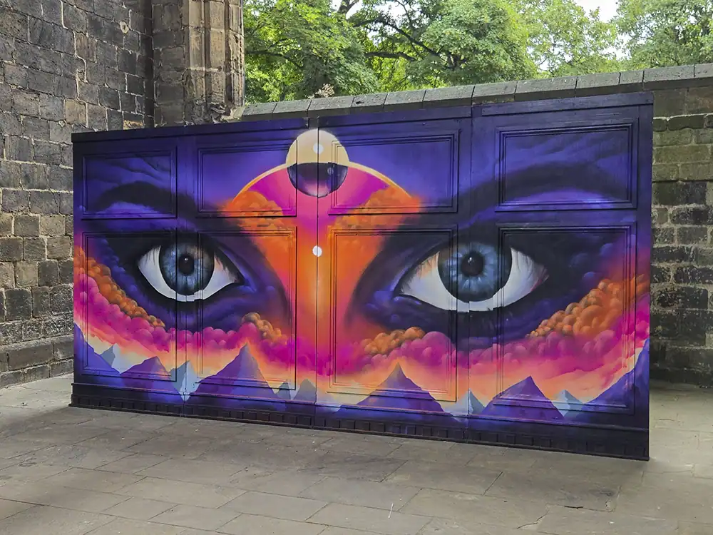 Four large wooden theatre flats hand-painted with striking blue eyes, a pale moon between them, and orange-pink clouds over purple mountains below.